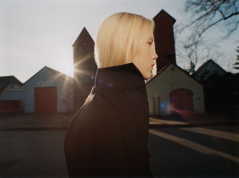 Profile portrait of a blonde model wearing a dark high-collared jacket outdoors against a street and building background at sunset.