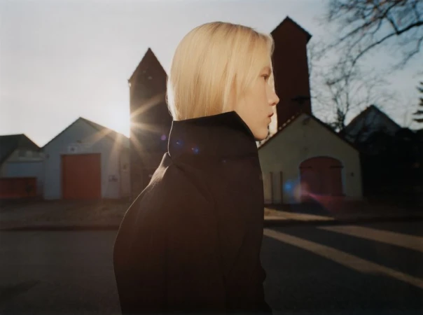 Profile portrait of a blonde model wearing a dark high-collared jacket outdoors against a street and building background at sunset.