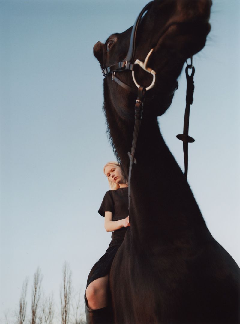 Low-angle shot of a black horse with a female model riding against a pale sky for Achtung Magazine.