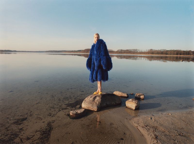 Model standing on a rock in shallow water wearing a voluminous blue coat and yellow shoes.