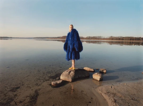 Model standing on a rock in shallow water wearing a voluminous blue coat and yellow shoes.