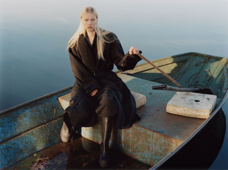 A blonde model wearing a dark coat and boots seated in a green wooden rowboat on a calm body of water.