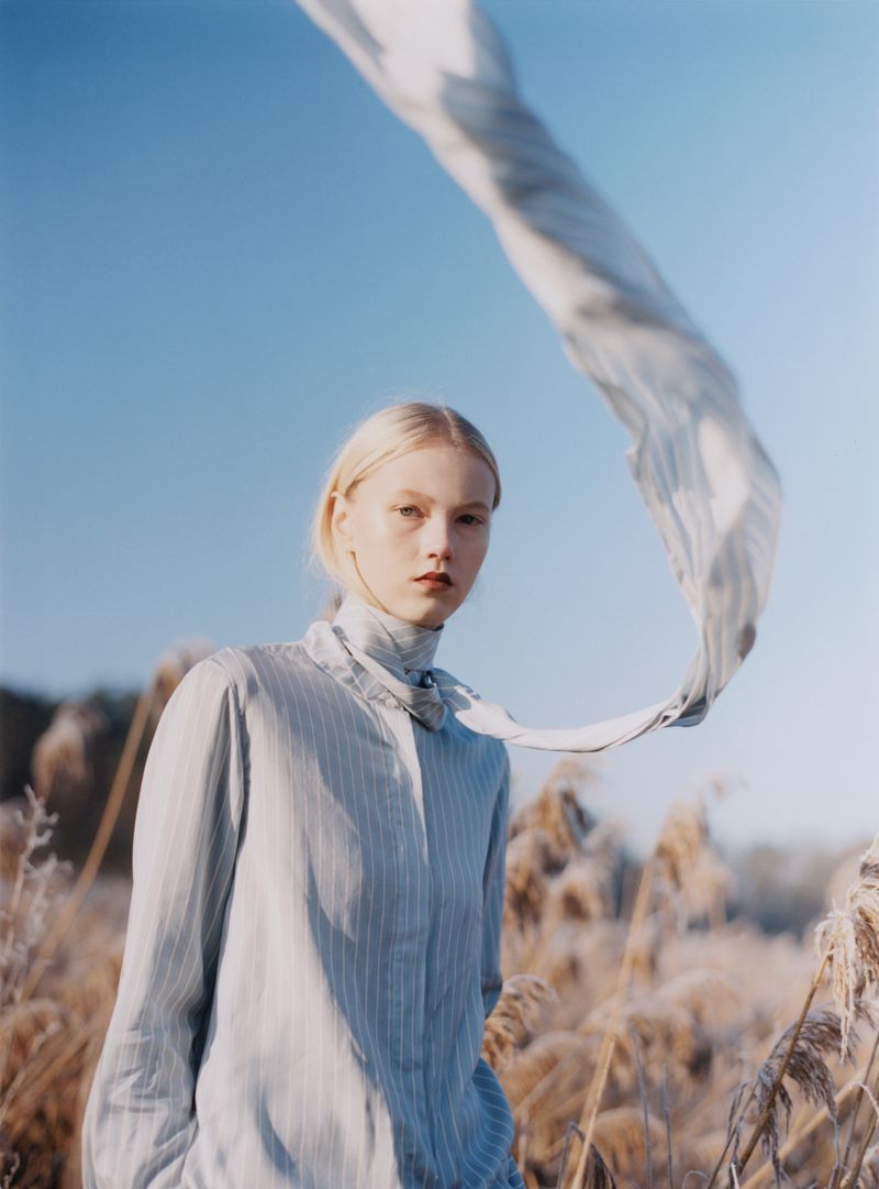 Blonde female model wearing a striped light blue shirt outdoors under a clear blue sky with a blowing scarf in Achtung Magazine editorial