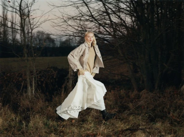 Model Malin Rudnick walking through a sparse woodland in a tan jacket and long white skirt for Achtung Magazine.