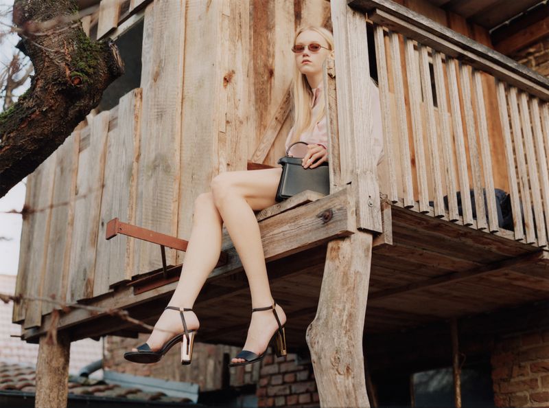 Model seated on a wooden balcony wearing high-heeled sandals holding a dark handbag for Achtung Magazine