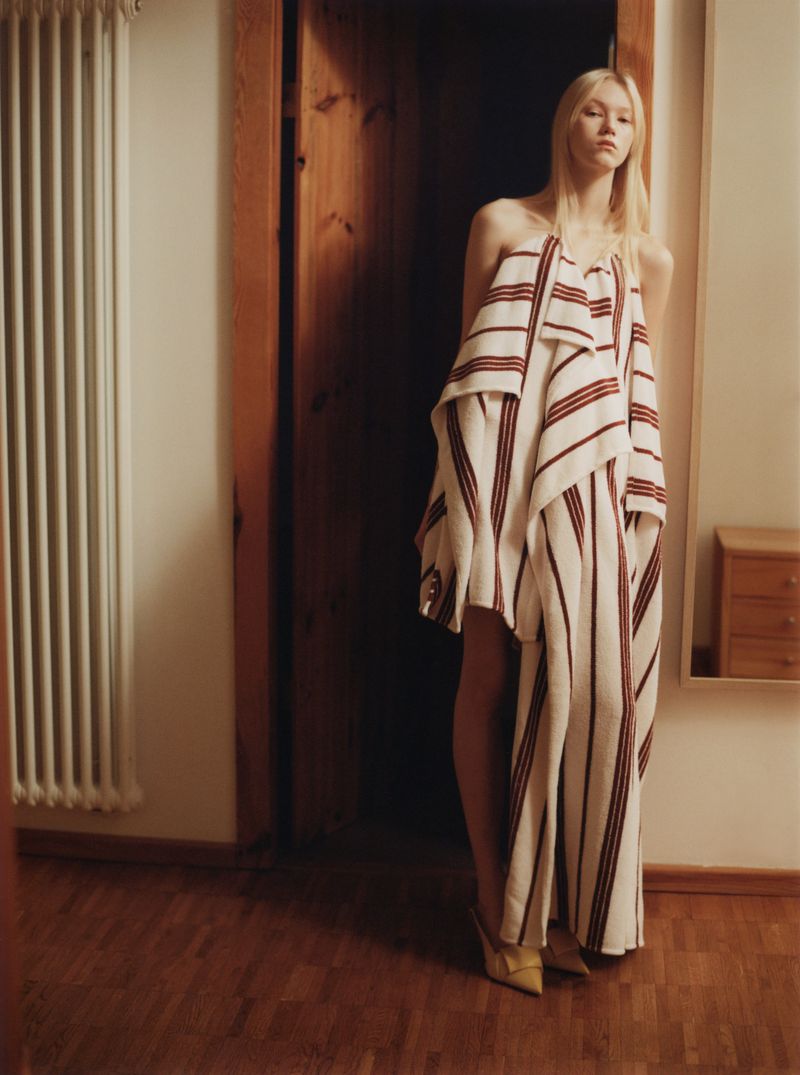 Model standing in a doorway wearing a draped white and red striped garment over wooden flooring.