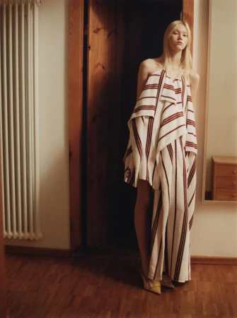 Model standing in a doorway wearing a draped white and red striped garment over wooden flooring.