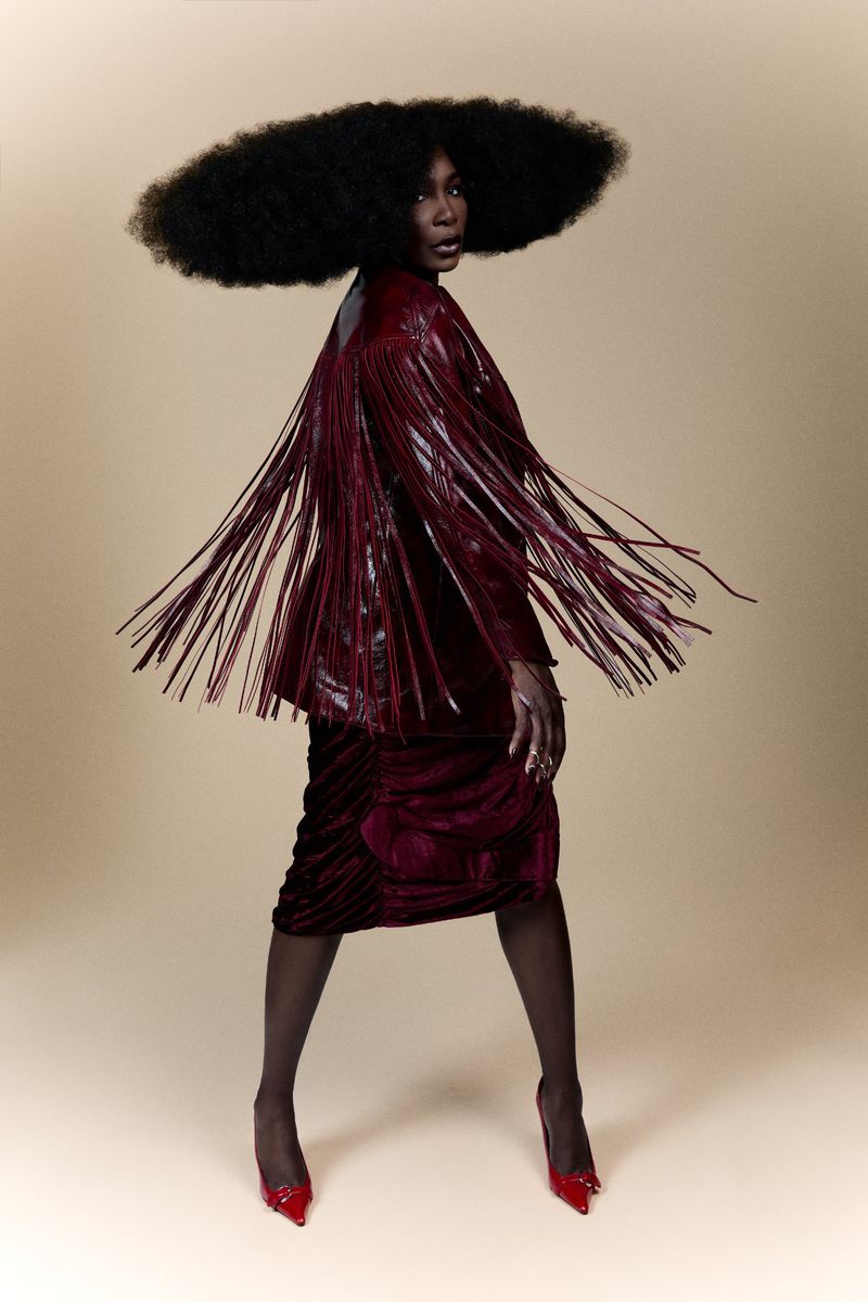An African American female fashion model poses in a studio wearing a red fringed jacket, red skirt, red heels and a large afro hairstyle.