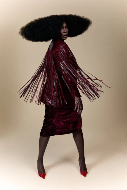 An African American female fashion model poses in a studio wearing a red fringed jacket, red skirt, red heels and a large afro hairstyle.
