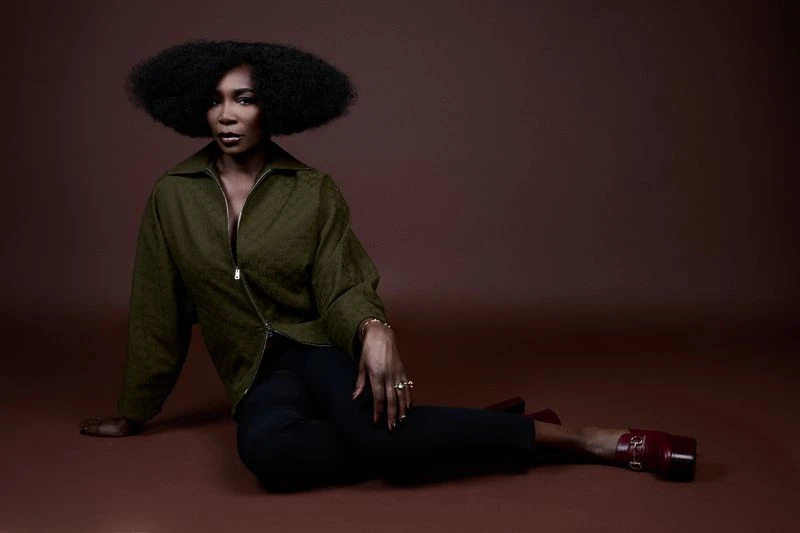 Portrait of African American woman with afro hairstyle wearing a green jacket and black pants, seated on a brown background.