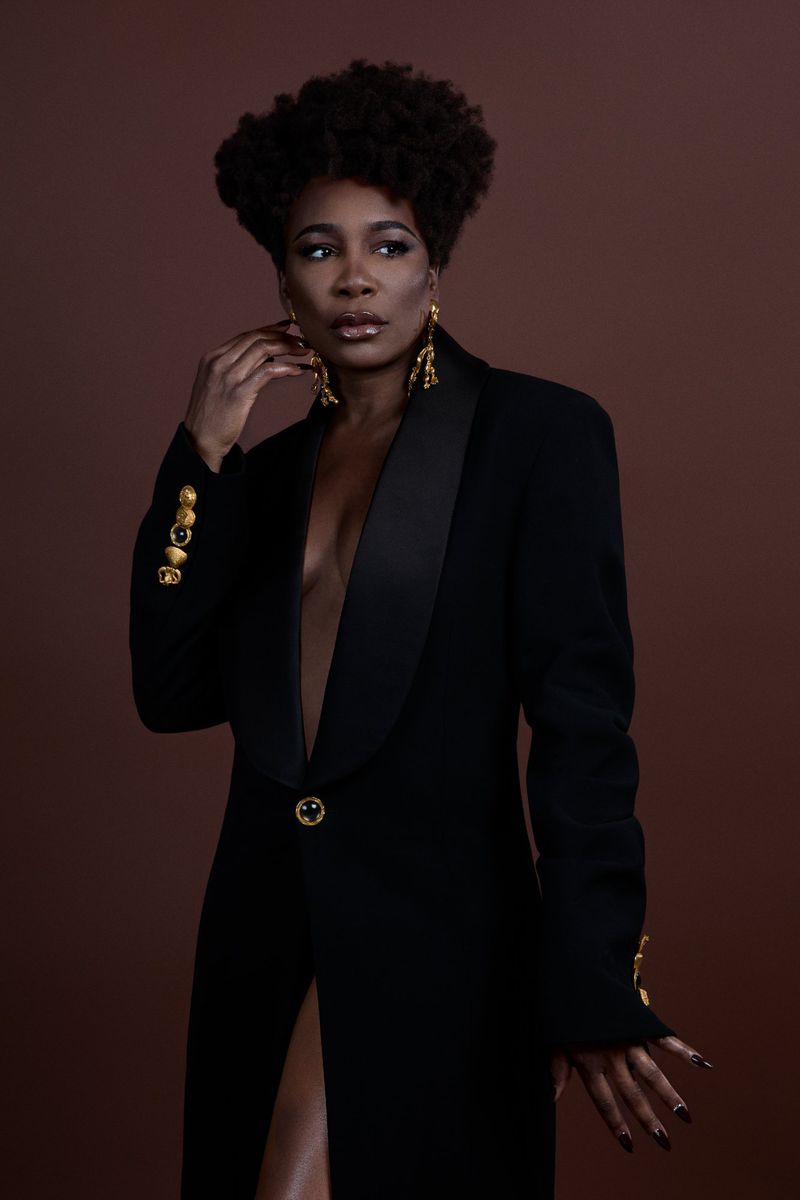 Glamorous portrait of an African-American woman in a black blazer and large gold earrings against a warm brown background.