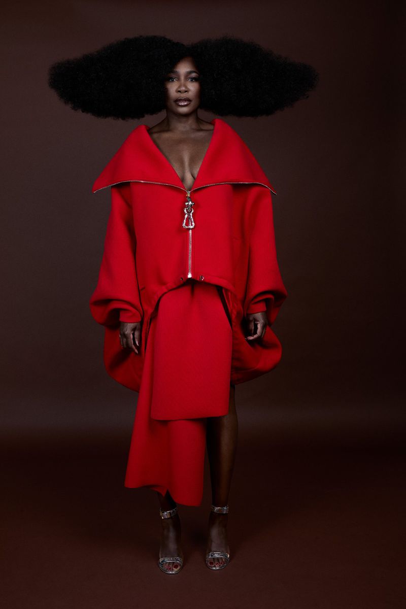 Woman with afro stands in a red asymmetrical dress and silver heels against a brown background in a fashion studio shot.