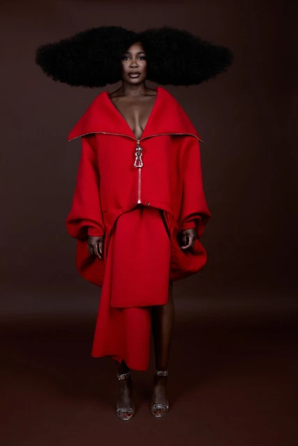 Woman with afro stands in a red asymmetrical dress and silver heels against a brown background in a fashion studio shot.