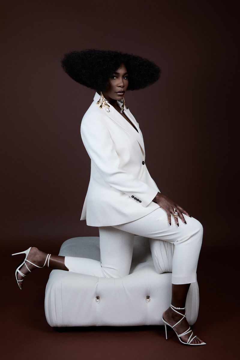 Stylish Black woman with afro in a white suit and strappy heels poses kneeling on a tufted ottoman against a brown background.