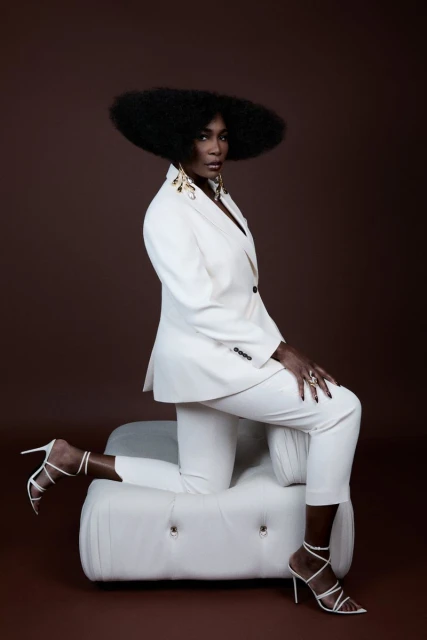 Stylish Black woman with afro in a white suit and strappy heels poses kneeling on a tufted ottoman against a brown background.