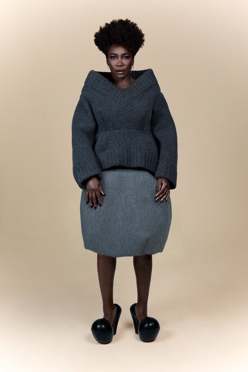 Stylish woman in gray sweater and sculptural skirt with sphere shoes poses in a studio with soft lighting and a neutral backdrop.