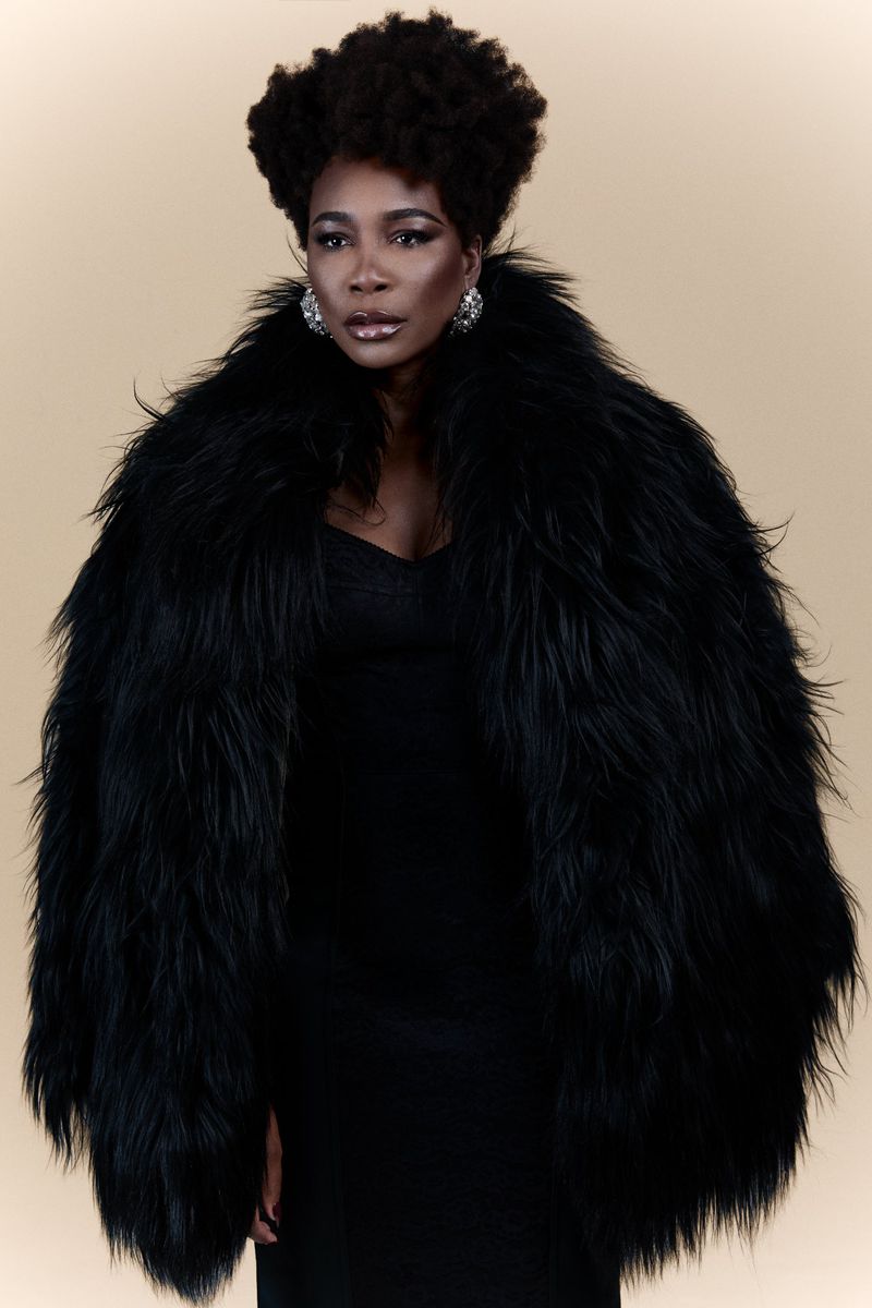 Portrait of a confident Black woman in a black dress and a large black faux fur coat, posing against a cream-colored background.