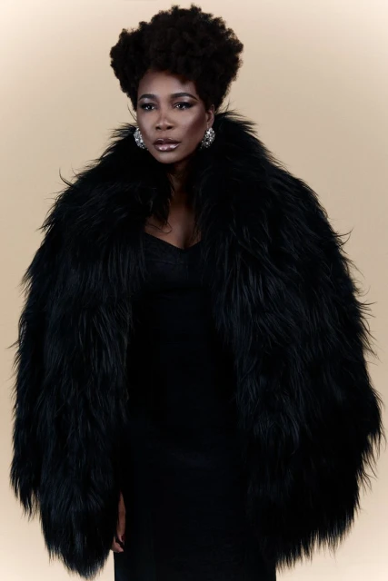 Portrait of a confident Black woman in a black dress and a large black faux fur coat, posing against a cream-colored background.