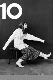 Model wearing an oversized white shirt and plaid trousers seated on a stool against a dark background with large number 10 text