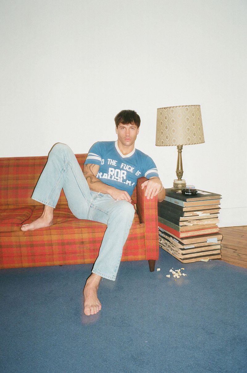 Man sitting barefoot on a plaid patterned sofa wearing a blue t-shirt and light wash jeans in an interior room.