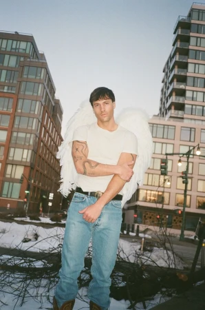 Man wearing large white angel wings, a white t-shirt, and blue jeans standing outdoors in front of tall city buildings.