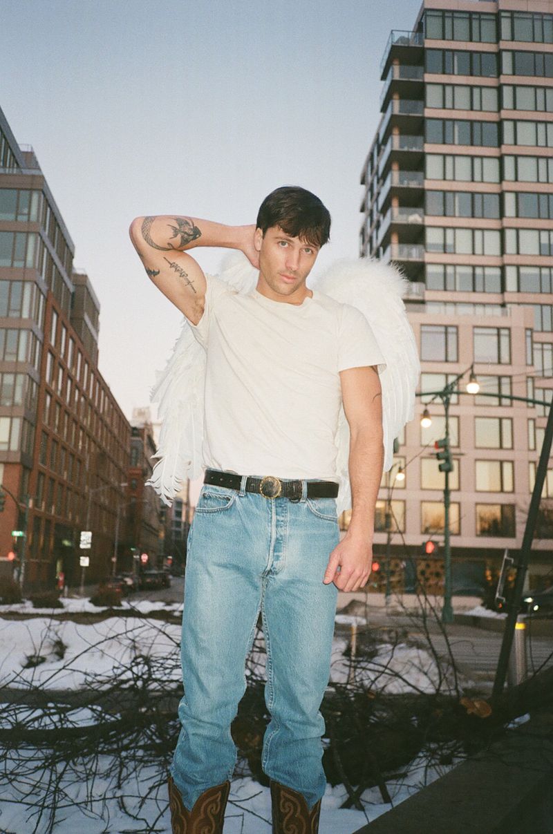 Man wearing angel wings, white t-shirt, and jeans standing on a city street for Interview Magazine.