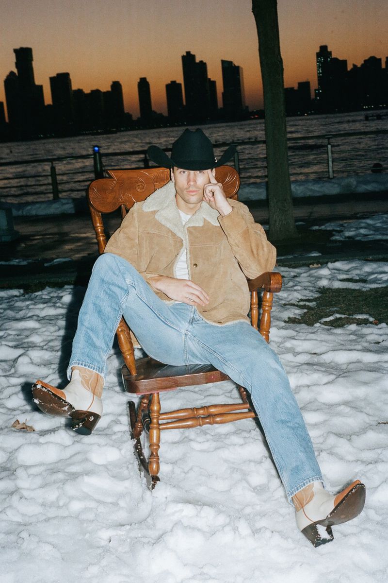 Man wearing a cowboy hat, shearling jacket, and blue jeans sitting on a wooden rocking chair outdoors in a snowy park at dusk.