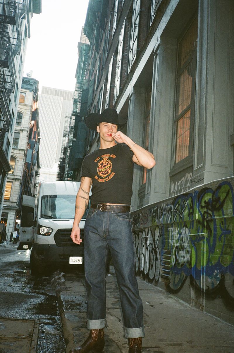 Man wearing a black cowboy hat and t-shirt standing on a city street beside a parked white van and graffiti wall