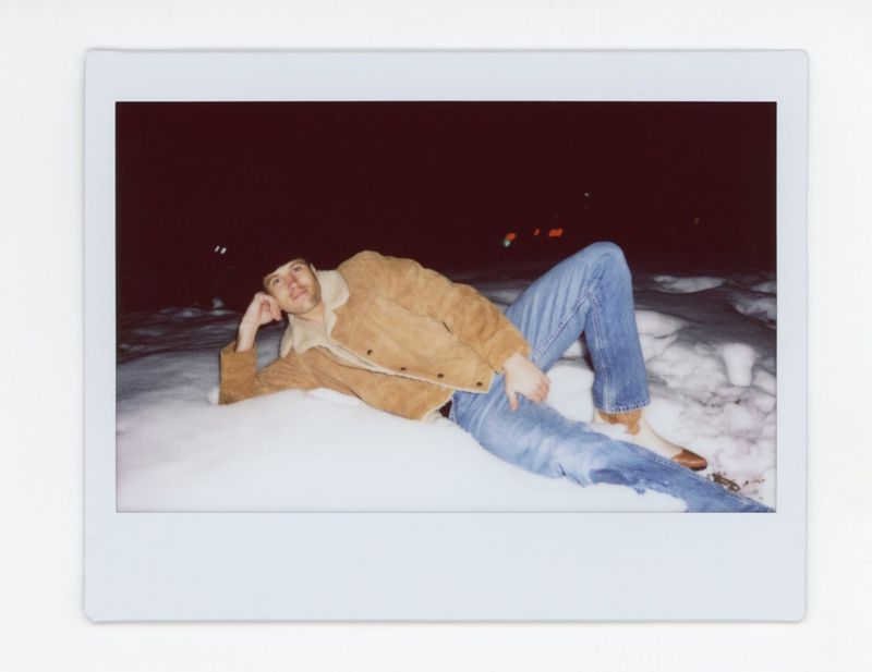 Man reclining in the snow wearing a tan shearling jacket and blue jeans, framed within a white polaroid border.