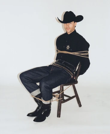 Man wearing a black cowboy hat and shirt tied to a chair with rope against a white seamless backdrop