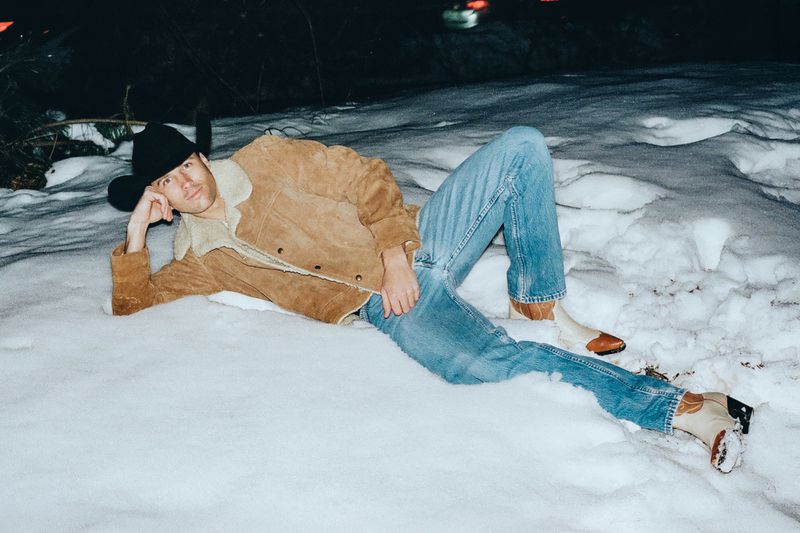 Man wearing a brown shearling jacket, cowboy hat, and blue jeans lying outdoors in the snow