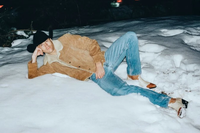 Man wearing a brown shearling jacket, cowboy hat, and blue jeans lying outdoors in the snow
