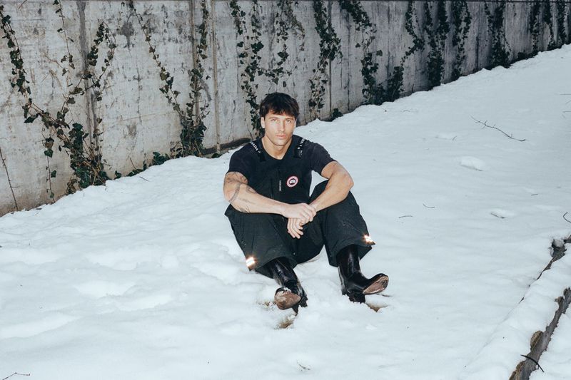 Man sitting on snow-covered ground wearing a black t-shirt and trousers with dark boots against a concrete wall