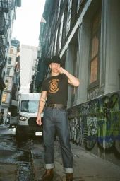 Man wearing a black cowboy hat and t-shirt standing on a city street beside a parked white van and graffiti wall