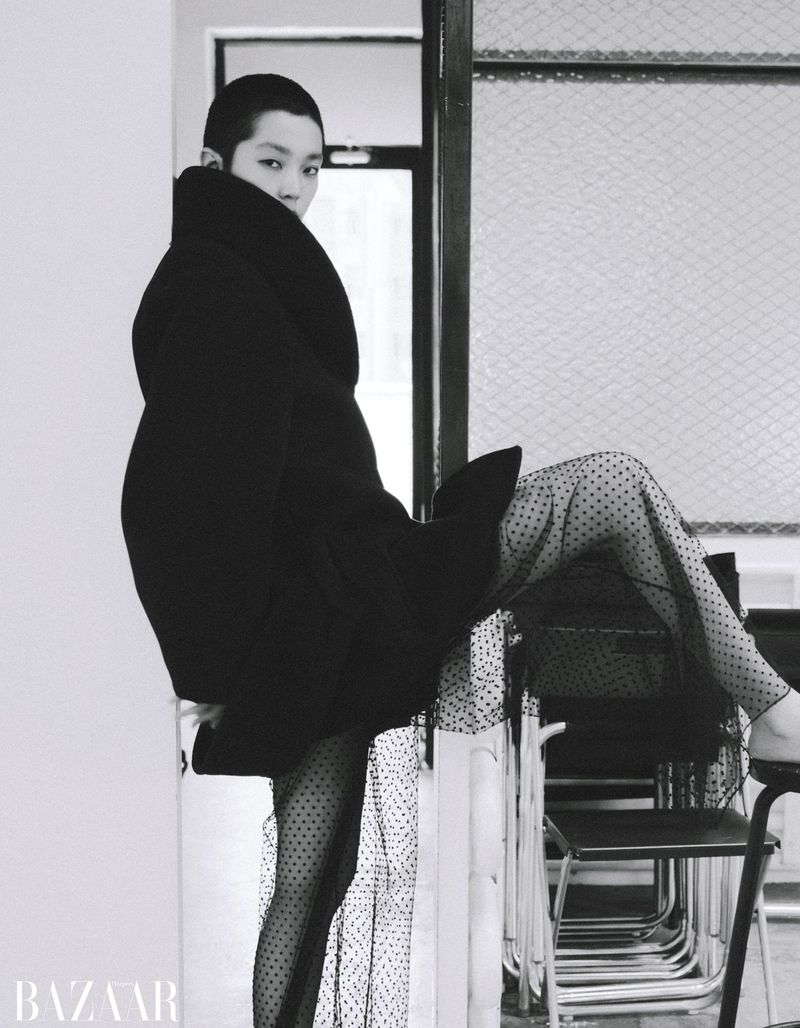 Model with a buzzcut wearing a heavy black coat and sheer polka dot stockings posing in a room
