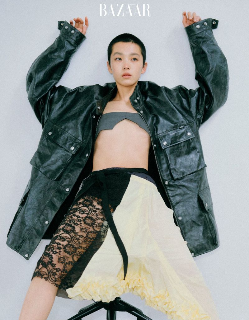 Model with a buzzcut wearing an oversized black leather jacket and a patterned lace skirt against a light grey background.