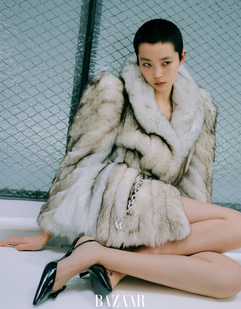 Model with a shaved head wearing a voluminous white fur coat and black heels against a wire mesh fence background