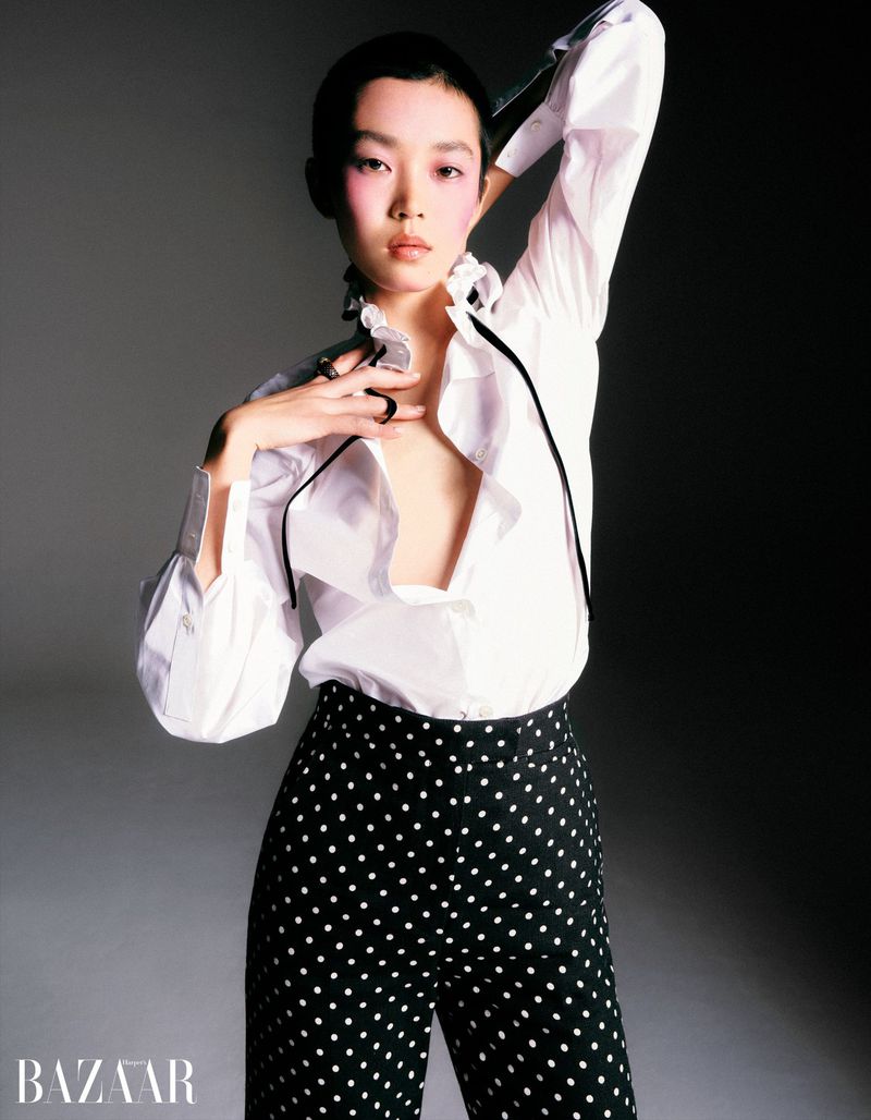 Model with short dark hair wearing a white ruffled shirt and black polka dot trousers against a grey backdrop for Harper's Bazaar China.