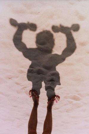 Overhead shot of a person standing on sand casting a shadow shaped like a muscular weightlifter holding dumbbells