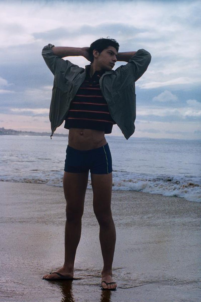 Model standing on a sandy beach wearing a striped shirt, green jacket, and dark swim trunks with hands behind head