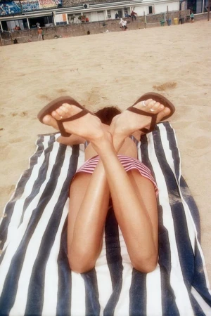 Close-up of crossed legs wearing flip-flops lying on a striped towel on a sandy beach