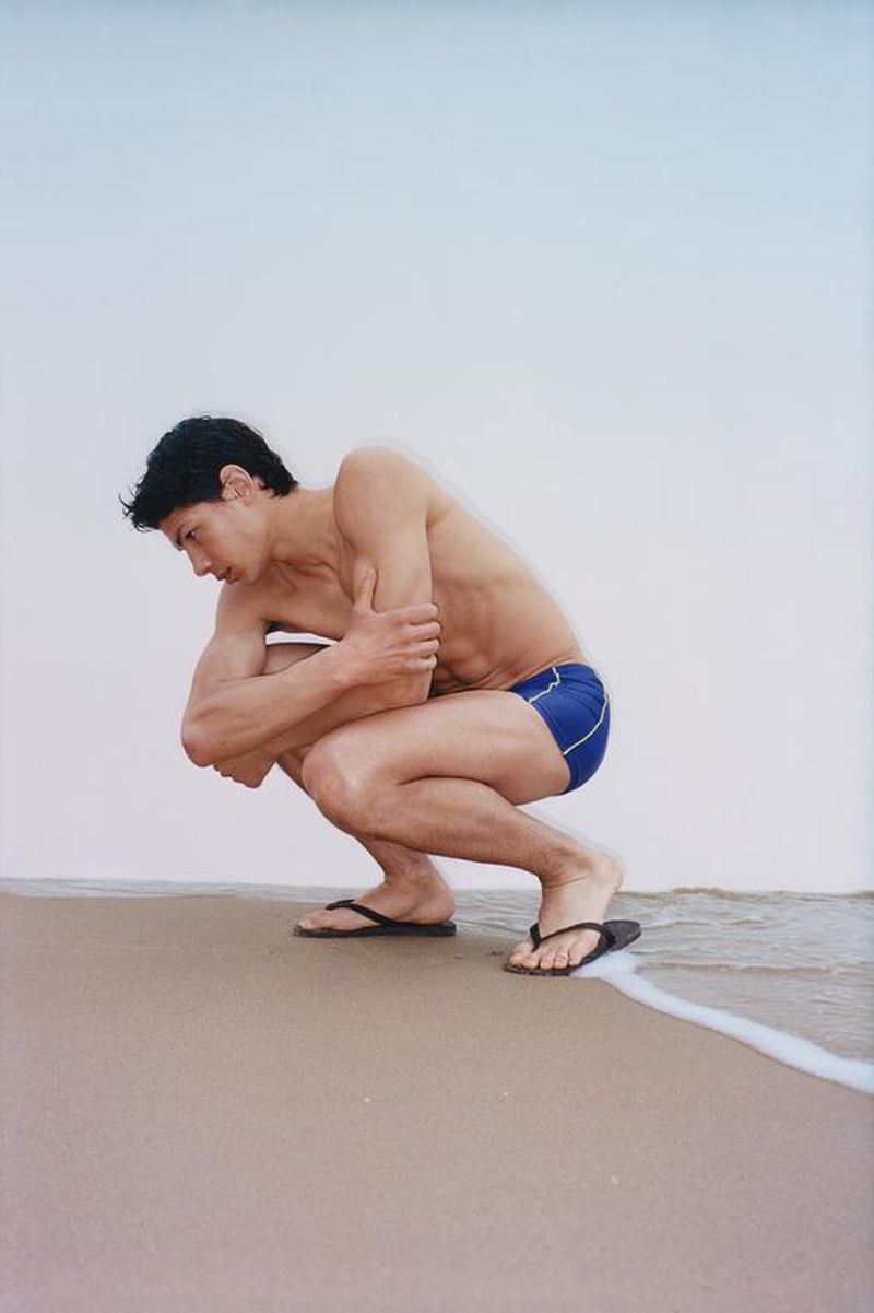 A man crouching in blue swim trunks and flip-flops on a wet sandy beach near gentle ocean waves against an overcast sky.
