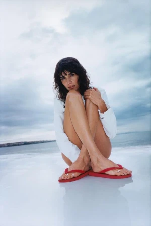 Model seated on a light reflective surface wearing white shirt and red flip flops under an overcast sky.