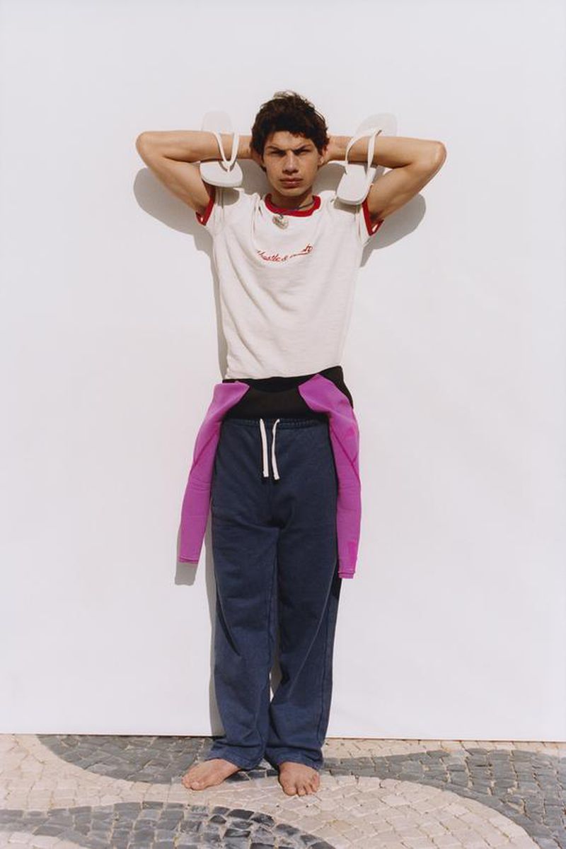 Model holding white flip-flops behind his neck, wearing a ringer t-shirt and dark trousers against a white backdrop.