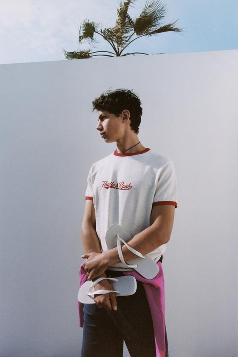 Young male model holding flip-flops wearing a white ringer t-shirt against a plain white wall with a palm tree top
