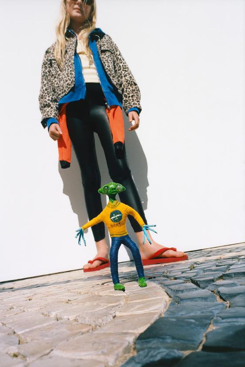 Young woman wearing jacket and black pants with an alien toy standing on a patterned stone surface near a white wall in an outdoor setting.