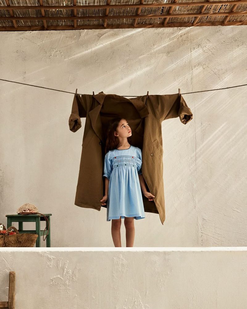 Young child in a blue smocked dress standing behind a brown coat hanging on a clothesline against a textured wall