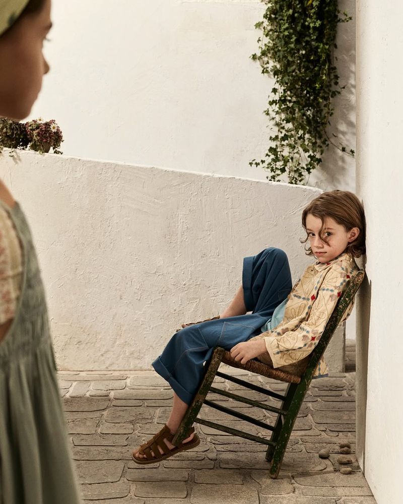 Child seated on a rustic green wooden chair leaning against a white wall with another child blurred in the foreground