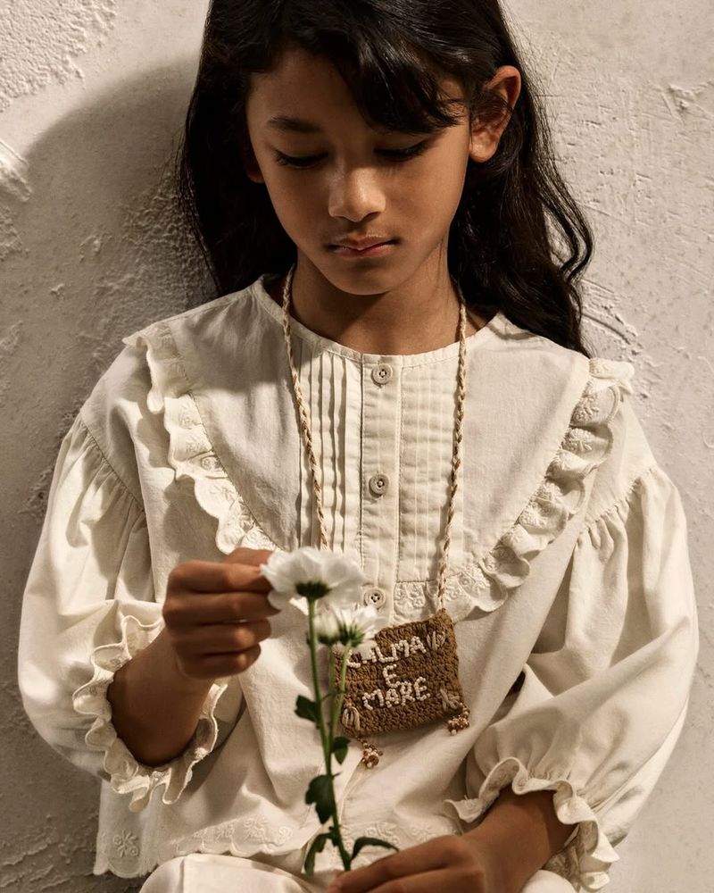 Young child wearing a cream ruffled blouse holding a white flower against a textured neutral wall for Zara Kids
