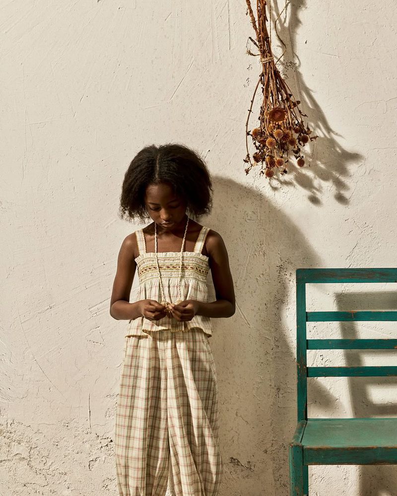 Young child in a plaid outfit standing beside a rustic teal chair against a textured beige wall with dried botanicals